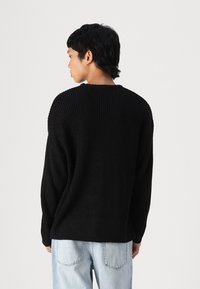 Suéter negro de punto acanalado con un corte relajado y cuello redondo, usado sobre unos vaqueros de mezclilla azul claro. Presenta mangas largas y una superficie texturizada.