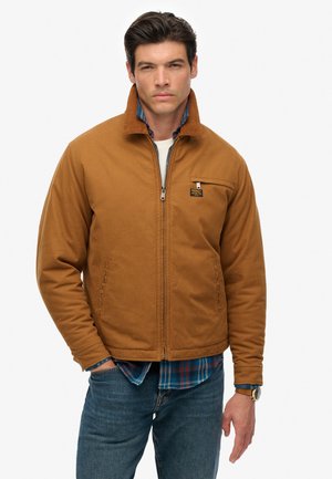 Man wearing brown zip-up jacket over blue plaid shirt and jeans, standing with one hand in pocket against a plain white background.