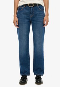 Jeans de denim azul con un diseño de pierna recta, trabillas y costuras en contraste. Combinados con zapatos negros y un cinturón a juego.