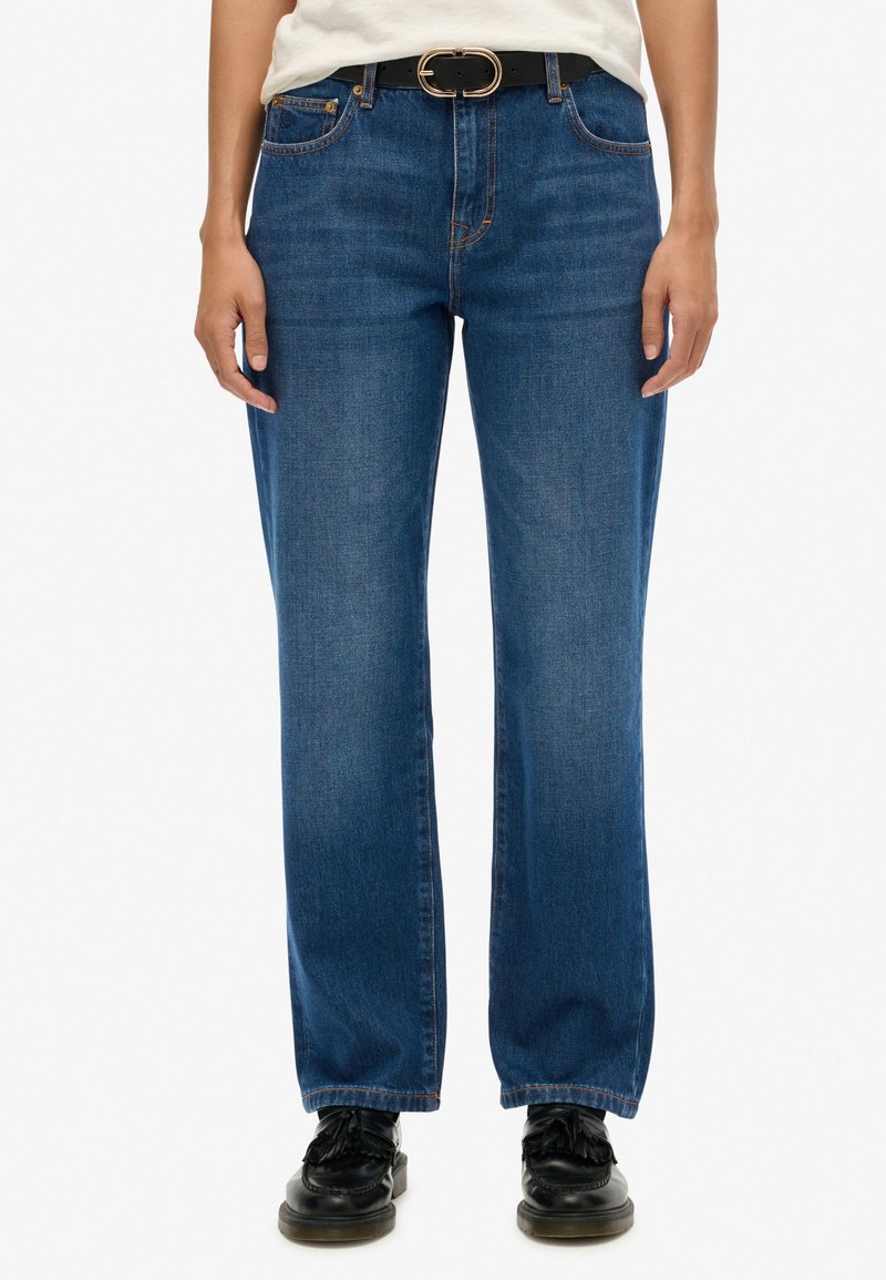 Jeans de denim azul con un diseño de pierna recta, trabillas y costuras en contraste. Combinados con zapatos negros y un cinturón a juego.