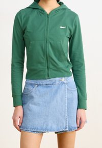 Grön hoodie med dragkedja i bomull, med framficka och vit logotyp. Matchas med en blå jeansminikjol med sidofickor och knappdetalj.