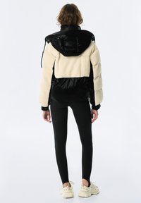 Czarna i kremowa kurtka puchowa o puszystej fakturze, z przerośniętym kapturem i elastycznymi mankietami, w połączeniu z czarnymi legginsami i jasnymi trampkami.