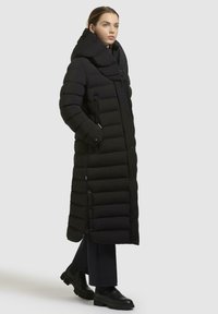 Lungo cappotto imbottito nero con cappuccio oversize, texture trapuntata, bottoni visibili e tasche con zip. Indossato sopra pantaloni scuri e stivali.