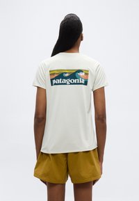 Άτομο με μακριά σκούρα μαλλιά που φοράει λευκό t-shirt Patagonia με γραφικό κύμα και μουσταρδί σορτς, γυρισμένο προς τα πίσω σε απλό λευκό φόντο.