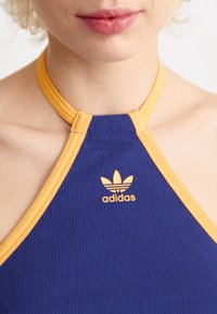 Adidas topp med halterneck i ribbat tyg, med blå kropp och gula accenter, samt en liten orange logotyp på framsidan.