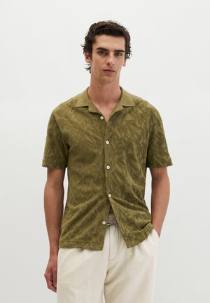Homme portant une chemise boutonnée à manches courtes à motifs vert olive et un pantalon beige clair, debout devant un fond blanc uni.