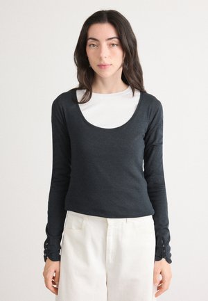 Donkergrijze long-sleeve top met een ronde hals en knopenaccenten op de mouwen, gecombineerd met witte high-waisted broeken. Zachte textuur.