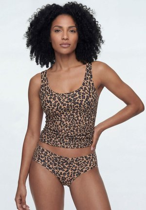 Femme aux cheveux bouclés portant un débardeur imprimé léopard et un slip assorti, posant avec une main sur la hanche sur un fond neutre.