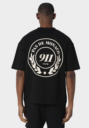 Mand iført sort oversize t-shirt med hvidt rundt emblem og teksten "PAS DE MONACO 911 CLUB" på ryggen, stående mod en ensfarvet baggrund.