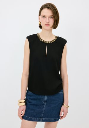 Top negro sin mangas con detalle de ojal, cuello adornado con acentos en oro y plata, combinado con una falda de mezclilla.
