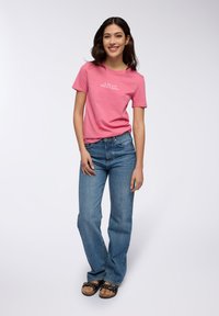 T-shirt di cotone rosa con maniche corte e testo bianco. Abbinata a jeans blu e sandali neri con borchie decorative. Design semplice.