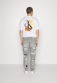 Mies, joka on selin, pukeutunut valkoiseen graafiseen T-paitaan ja harmaisiin revenneisiin farkkuihin, sekä valkoisiin lenkkareihin, tasaisella vaalealla taustalla.