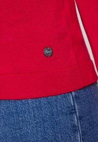 Roter gestrickter Pullover mit geripptem Saum, versehen mit einem schwarzen runden Esprit-Tag, der an der Seite genäht ist. Getragen über blauer Denim-Stoff.