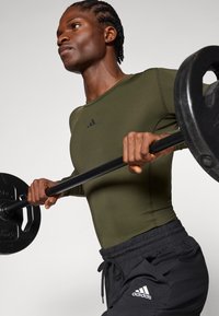 Tmavě zelené sportovní tričko s dlouhým rukávem a přiléhavým střihem, černé logo Adidas, a černé kalhoty s šňůrkou a bílým logem, držící činku.