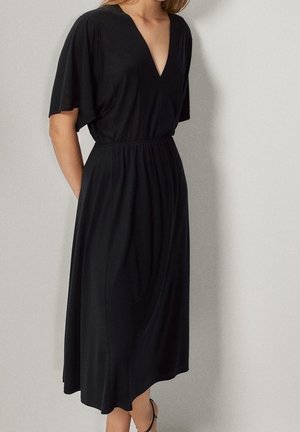 Femme portant une robe midi noire avec des manches courtes évasées, un décolleté en V, une taille cintrée et un ourlet asymétrique, debout contre un mur uni.