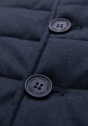 Close-up van donkerblauwe stof met twee ronde zwarte knopen, waarvan één vastgenaaid en gegraveerd met het woord "OMBRE" herhaald rond de rand.