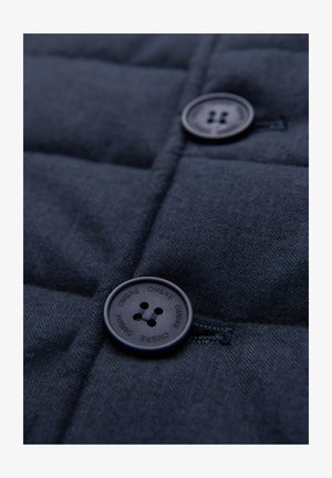 Close-up van donkerblauwe stof met twee ronde zwarte knopen, waarvan één vastgenaaid en gegraveerd met het woord "OMBRE" herhaald rond de rand.