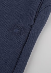 Marineblaue gestrickte Sweater mit gerippter Textur, der ein rundes Logo auf dem Ärmel trägt. Der Stoff scheint weich und dehnbar zu sein.