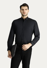 Camisa negra de manga larga con cuello abotonado, hecha de una tela suave; combinada con jeans negros ajustados y sosteniendo un bolso de mano texturizado negro.