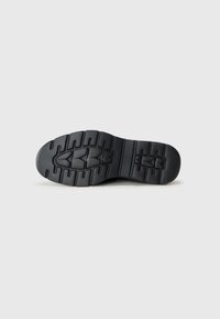 Black rubber shoe sole featuring a textured tread pattern with grooves for traction. Rounded toe and flat design for stability.