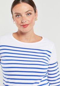 Witte blouse met lange mouwen en horizontale blauwe strepen. Ronde halslijn en een zachte, gladde textuur. Heeft een logo op de mouw.