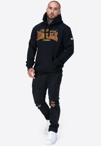 Felpa nera in cotone con tasca a marsupio frontale, caratterizzata da un grande logo giallo e rosso. Abbinata a jeans neri strappati e sneakers bianche.