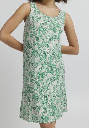 Vestido blanco sin mangas con patrón floral verde, llevado por una persona con los brazos doblados detrás de la espalda, mostrado desde el cuello hasta las rodillas.
