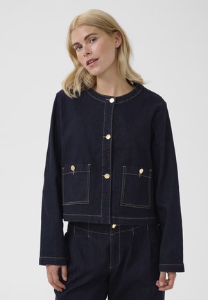 Blonde Frau trägt eine dunkelblaue Jeansjacke mit goldenen Knöpfen und zwei vorderen Taschen, kombiniert mit passenden Jeanshosen.