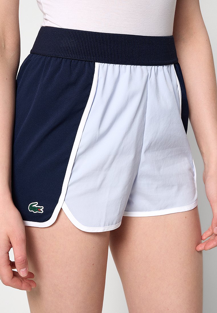 Shorts de sport bleus marines et blancs avec une texture en maille, une taille élastique et des fentes sur les côtés, ornés d'un petit logo crocodile vert.