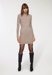 Abito a maniche lunghe color beige con collo a imbuto e silhouette aderente, abbinato a stivali neri lucidi al ginocchio. Texture liscia in tutto l'abito.