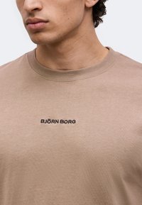Gros plan d'une personne portant un t-shirt beige avec "BJÖRN BORG" brodé en noir sur la poitrine.