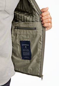 Interior de chaqueta acolchada color verde oliva con un bolsillo con cremallera, etiqueta que detalla la sostenibilidad del tejido y textura suave. Cierre con cremallera en el borde.