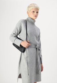 Siva turtleneck obleka iz mehkega materiala s pasom in stranskim razporkom, v kombinaciji z majhno črno torbico in prilegajočimi rokavi.