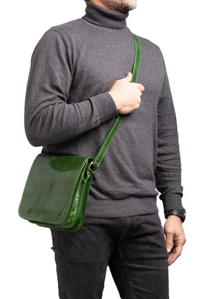 Homme portant un col roulé gris foncé et un jean noir, tenant la sangle d'un sac bandoulière en cuir vert sur l'épaule.