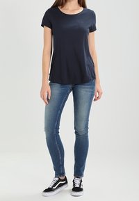 Žena v námořnickém modrém tričku s krátkým rukávem, modrých skinny džínách a černo-bílých teniskách, stojící na jednolité světlé pozadí.