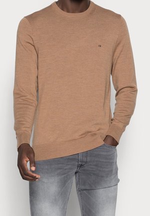 Mand iført en lys brun langærmet sweater med "ck" logo på brystet og lysegrå jeans, hånd i lommen.