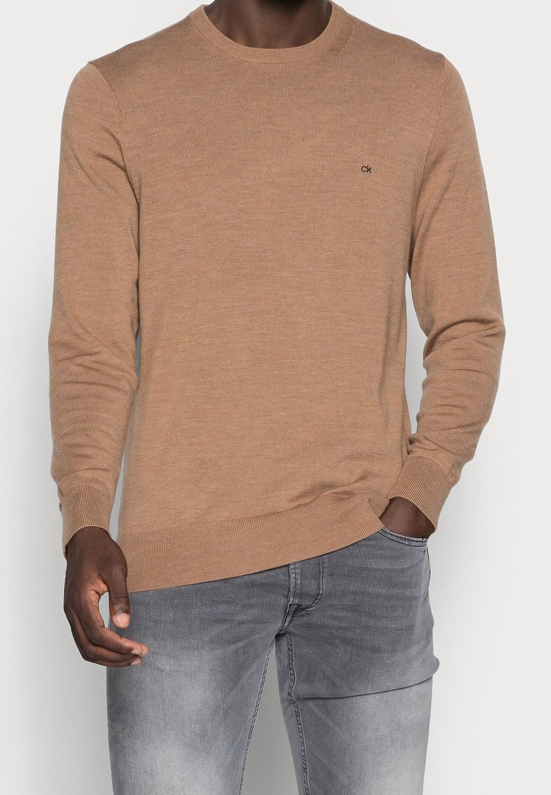 Homme portant un pull marron clair à manches longues avec le logo "ck" sur la poitrine et un jean gris clair, main dans la poche.