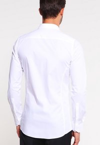 Camisa blanca de manga larga hecha de tejido suave. Presenta un cuello puntiagudo, puños con botón y un corte ajustado con costuras en la parte posterior para dar forma.