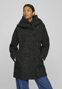 Manteau noir texturé avec un grand col replié, fermeture à un bouton, et poches latérales, porté sur une tenue en denim bleu.