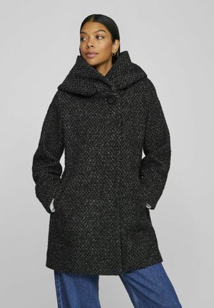 Černý texturovaný kabát s velkým skladaným límcem, uzavíráním na jeden knoflík a bočními kapsami, nošený přes modrý denimový outfit.