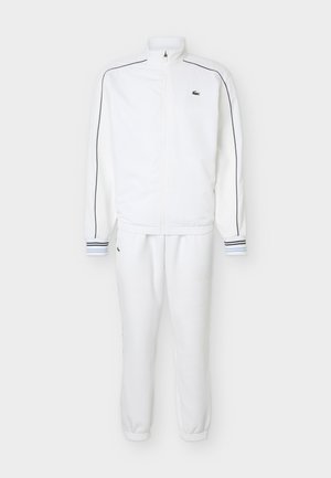 Λευκή φόρμα με φερμουάρ και μαύρη ρίγα στα μανίκια, ριμπ μανσέτες με ρίγες και κεντητό πράσινο λογότυπο κροκοδείλου στο στήθος και το παντελόνι.