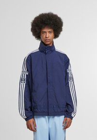 Námornícky modrá bunda adidas s vysokým golierom, biele pruhy pozdĺž rukávov a nášivka loga na ľavom rukáve. Hladká, ľahká tkanina.