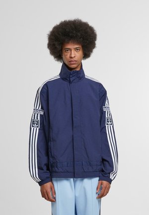 Navy blue adidas jacket with high collar, white stripes along sleeves, and logo patch on the left arm. Smooth, lightweight fabric.