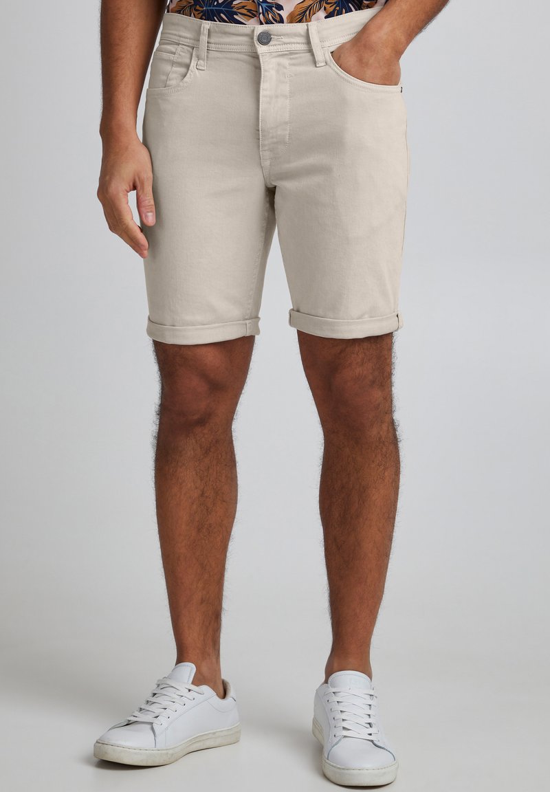 Shorts in denim beige con orli arrotolati, cinque tasche, chiusura con bottone in metallo e design aderente, abbinati a sneakers bianche.