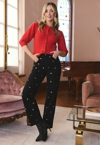 Femeie stand într-o cameră purtând un top tricotat roșu cu fundițe, pantaloni negri cu inimioare aurii și cizme negre cu toc, lângă mobilă vintage și o fereastră din sticlă colorată.