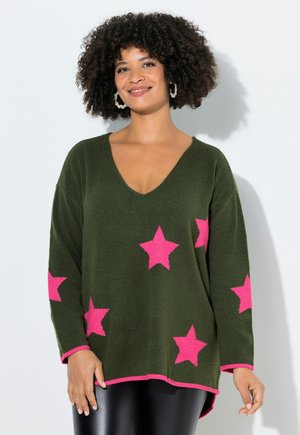 Kvinde med krøllet hår, smilende, iført mørkegrøn V-hals sweater med lyserøde stjerner og sorte bukser mod hvid baggrund.