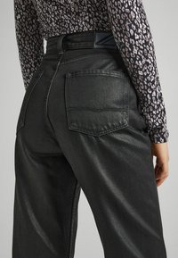 Jeans de mezclilla negra con una textura elegante, que cuentan con dos bolsillos traseros y una cinturilla con marca. La tela tiene un ligero brillo.