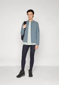 Casaco azul-claro com fecho de correr por cima de uma t-shirt verde, combinado com leggings pretas com painéis contrastantes e botas de caminhada pretas com detalhes em verde.