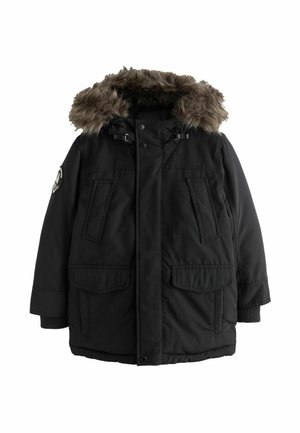 Junge mit schwarzem Wintermantel mit pelzbesetzter Kapuze und blauer Jeans steht vor einem schlichten hellen Hintergrund.