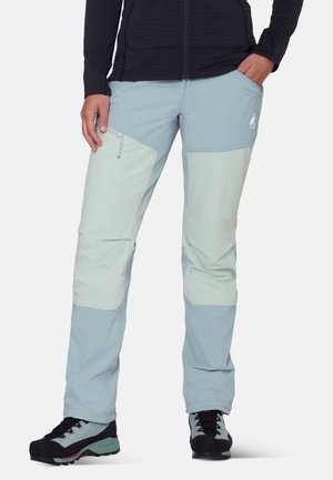 Vrouw die tweekleurige lichtblauwe wandelbroek draagt met een afritsbare dijbeenzak en zwarte wandelschoenen, handen in de zakken, staand tegen een witte achtergrond.